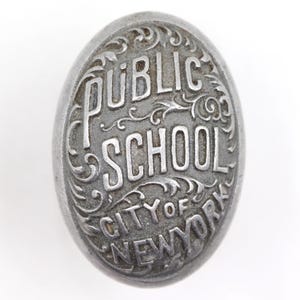 Op de afbeelding: Ovale, antieke, zilverkleurige metalen badge. De badge heeft de woorden "PUBLIC SCHOOL CITY OF NEW YORK" in verhoogde letters met decoratieve krullen. Het ontwerp staat tegen een witte achtergrond.