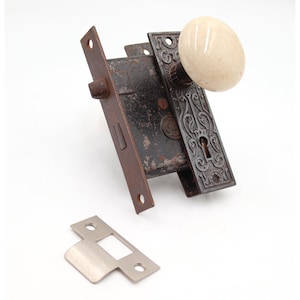 May include: Vintage door hardware set. The set includes a dark, ornate metal lock plate with a cream-coloured oval doorknob. The set has a matching strike plate and a rusty, aged appearance, suggesting vintage charm.