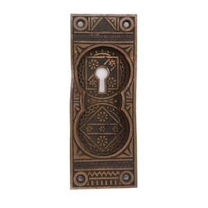 Może przedstawiać: Antyczna osłona otworu na klucz wykonana z mosiądzu z misternym geometrycznym wzorem. Osłona jest prostokątna i ma otwór na klucz w środku. Wzór jest w ciemnobrązowym kolorze z złotym wykończeniem.