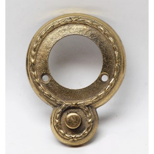 May include: A gold-toned metal doorbell button with a decorative wreath design. The button is round and has a smaller, round button in the center.