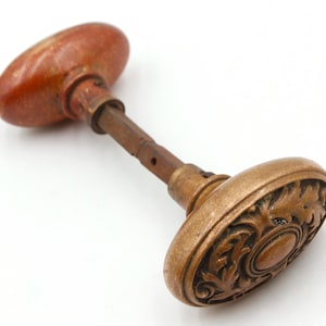 May include: Antique bronze door knob with an oval shape and ornate leaf design. The knob has a reddish-brown finish on one side and a bronze finish on the other. The connecting rod is a dark brown color.