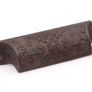 May include: A decorative, rectangular, brown metal drawer pull with an ornate, textured surface. The surface features a repeating pattern of circles, lines, and floral-like shapes. The ends have holes for mounting.