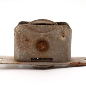 May include: A worn metal roller assembly, showing signs of rust and age. It features a rectangular housing with a circular wheel and a flat base with mounting holes. The metal has a weathered appearance.