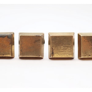 May include: Four square, brass-colored metal objects are arranged horizontally against a white background. Each object has a slightly weathered appearance, with variations in color and texture. The objects appear to be hardware or decorative elements.