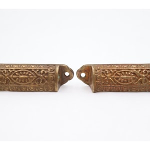 Op de afbeelding: Twee antieke messing lade-grepen met sierlijke bloemen- en krulwerk ontwerpen. Elke greep heeft een rechthoekig lichaam, een gebogen handvat en twee montagegaten. Het messing heeft een warme, verouderde patina.