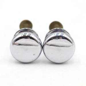 Puede incluir: Dos pomos de armario cromados con una superficie brillante y reflectante. Los pomos tienen forma redondeada y están montados sobre una base de color latón. El herraje se encuentra sobre un fondo blanco liso.