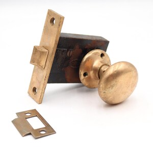 May include: A brass door knob set featuring a round handle, a rectangular faceplate with two screw holes, and a dark metal internal latch mechanism. A separate small rectangular brass strike plate with two screw holes is positioned in front.
