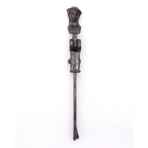 May include: A vintage cast iron door knocker in the shape of a person wearing a hat. The knocker is black with rust spots and has a long, thin handle.