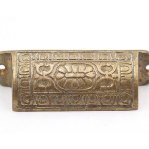 May include: A vintage brass drawer pull with an ornate floral design. The rectangular handle features a detailed embossed pattern with a central flower and symmetrical scrollwork. The pull has two mounting holes on each end.