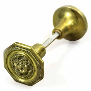 May include: Antique brass door knob with an octagonal base and a floral design. The knob has a round handle and a connecting rod. The brass has a slightly aged appearance, suggesting vintage or antique status.