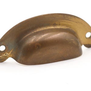 May include: A vintage-style, solid brass drawer pull with a curved, cup-like design. The hardware has two holes for mounting and shows signs of age and wear, with a warm, aged patina.