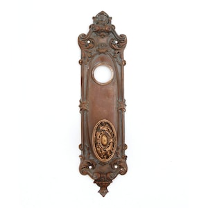 May include: Ornate antique brass door plate with a keyhole and decorative details. The plate has a dark brown patina and a raised design.