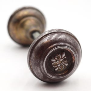May include: Close-up of two antique door knobs. The front knob is a dark, aged bronze color with a decorative floral design in the center. The second knob is out of focus, with a similar design and color.