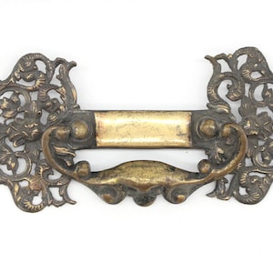 May include: Ornate brass drawer pull with a decorative floral design. The pull has a rectangular plate and a curved handle.