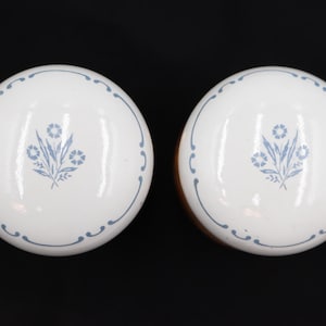 Pode incluir: Dois puxadores de armário de cerâmica branca com um desenho floral azul e uma borda decorativa azul. Os puxadores são redondos e têm um acabamento brilhante. O fundo é preto.