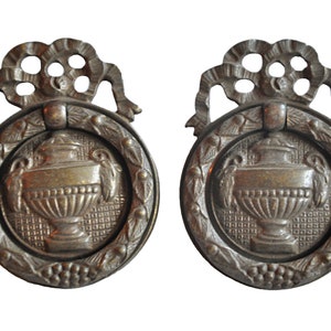 Puede incluir: Dos tiradores de puerta de armario de latón antiguo con un diseño decorativo que presenta una urna estilizada con una corona y un lazo.