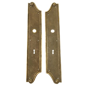 May include: Two antique brass door plates with a rectangular shape and decorative scrollwork at the top and bottom. Each plate has a keyhole and a circular hole for a doorknob. The plates are a warm, aged brass color.
