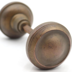 May include: A round, antique-style brass door knob with a weathered, darkened patina. The hardware features a smooth, circular face and a central spindle connecting to a matching circular backplate, showcasing a worn metallic finish throughout.