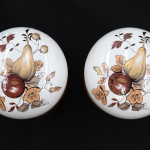 May include: Two white ceramic cabinet knobs with a fruit design. Each knob features a pear and an apple, with brown, gold, and orange floral accents. The knobs are round and have a glossy finish.