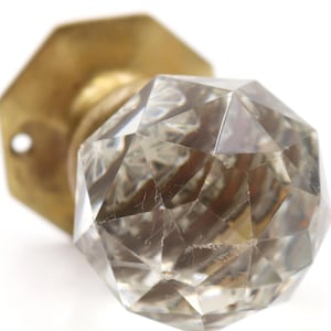 May include: A close-up shot of a vintage door knob featuring a faceted crystal handle and a brass base. The crystal is clear with internal imperfections, and the brass has a warm, aged patina. The knob is set against a white background.