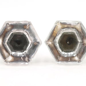 Può includere: Due portacandele esagonali in vetro trasparente. I portacandele hanno un interno scuro e un esterno color argento. I portacandele sono vuoti e poggiano su una superficie bianca. I portacandele sono uno accanto all'altro.