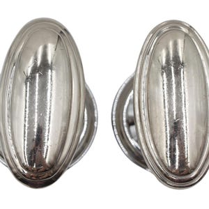 Op de afbeelding: Twee zilverkleurige ovale kastknoppen. Elke knop heeft een verhoogd, ovaal midden met een geribbelde rand, gemonteerd op een cirkelvormige basis. De knoppen zijn reflecterend en tonen licht en schaduw.