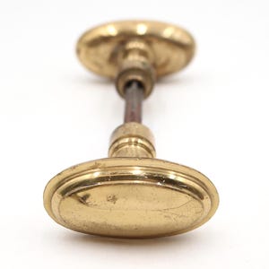 Può includere: Pomello per porta antico in ottone di forma ovale. Il pomello ha una finitura ossidata, che mostra segni di età. L'asta centrale è di colore marrone scuro. Il pomello è posto su uno sfondo bianco.