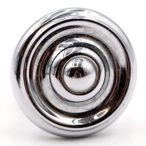 May include: A close-up of a shiny, chrome-finished cabinet knob. The knob has a circular design with concentric rings and a central, rounded button. The reflective surface creates highlights and shadows.