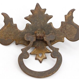 Op de afbeelding: Een vintage, sierlijke, metalen ladegreep met een decoratief, bladvormig ontwerp. De greep is roestbruin en heeft een ringvormige handgreep.