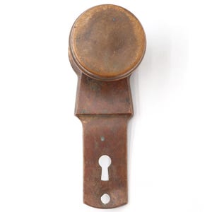 May include: Antique bronze door knob with a round handle and a rectangular backplate. The knob has a keyhole and a small hole at the bottom. The hardware is a warm brown color.