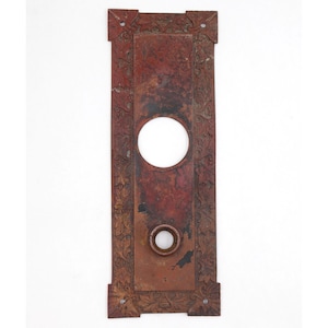 May include: A rectangular, antique door plate with a distressed, reddish-brown finish. It features a large circular opening and a smaller one below, with ornate floral detailing along the edges and corners. The plate is made of metal.