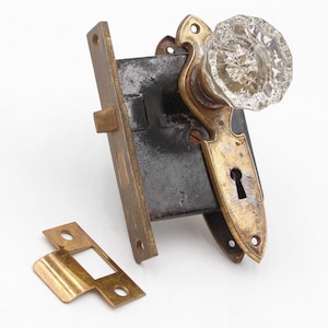 May include: Vintage door hardware set with a clear glass doorknob, brass-coloured escutcheon with a keyhole, and a black metal lock mechanism. Includes a separate brass-coloured strike plate. Suitable for restoration.