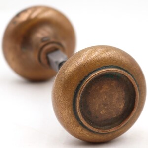 Peut inclure: Gros plan sur deux boutons de porte anciens en laiton. Les boutons sont ronds avec une surface légèrement texturée et une patine chaude et vieillie. La quincaillerie est posée sur un fond blanc, soulignant les détails du design vintage.