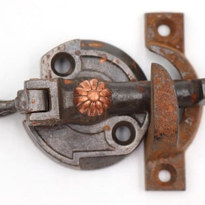 Pode incluir: Close-up de uma tranca de metal vintage com um detalhe em forma de flor. A tranca é uma mistura de cinza e marrom, com ferrugem visível. O design inclui uma base circular, um braço giratório e um fecho. A tranca mede cerca de 10 cm de comprimento.