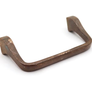 May include: A vintage, rectangular drawer pull with a curved handle. The metal hardware has a weathered, bronze finish with visible wear and patina. The pull is set against a white background.