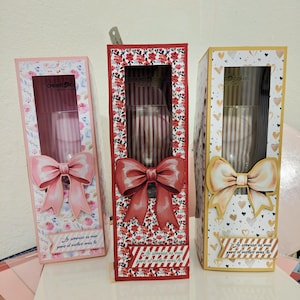 Puede incluir: Tres cajas de regalo decorativas con ventanas transparentes y lazos grandes. Las cajas son rosas, rojas y amarillas, con estampados florales y de corazón. El texto en las cajas dice "La sonrisa es mia pero el motivo eres tu."