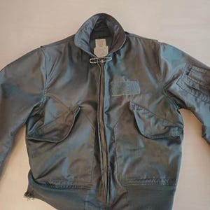 Op de afbeelding: Een vintage olijfgroene bomberjack met geribde zoom en manchetten. De jas heeft twee voorzakken, een patch op de borst en een ritssluiting. Een veiligheidsspeld zet de kraag vast.