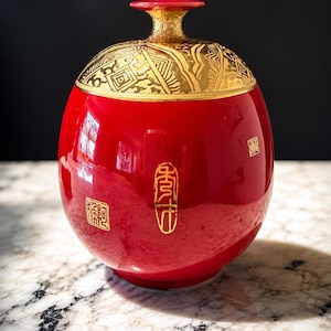 May include: A red, round ceramic jar with a gold-colored lid and accents. The lid features a geometric pattern. Gold Chinese characters are on the jar's surface. The jar has a glossy finish and is likely decorative.