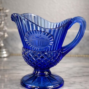 May include: A cobalt blue glass creamer with a decorative design. The creamer features a diamond pattern on the base and a detailed embossed image. The handle is curved, and the rim has a wavy design. A classic piece for serving.