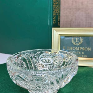 May include: A clear, decorative glass bowl with an intricate, embossed pattern. The bowl has a rounded shape and sits on a small base. The background includes a green book and a framed sign with the text "E. Thompson Estate & Suites."