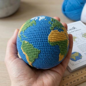 Könnte beinhalten: Ein gehäkelter Erdglobus, gehalten in einer Hand, mit grünen und gelben Kontinenten auf einem blauen Ozean. Das Wort "Earth" ist auf einem Papier zu sehen, was auf ein Bastelprojekt hindeutet. Ein Lineal und blaue Wolle sind ebenfalls im Hintergrund.