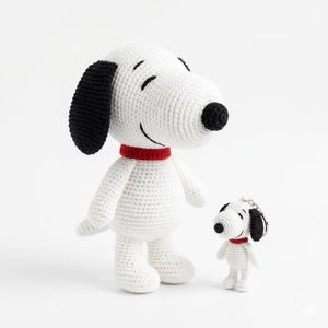 May include: A white crocheted Snoopy plush toy with black ears and nose, and a red collar. A smaller keychain version of Snoopy is also present. The toys are standing on a white surface.