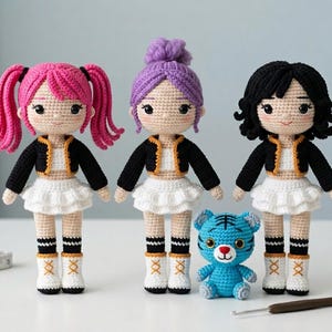 May include: Three handmade crochet dolls with pink, purple, and black hair, wearing black jackets, white skirts, and boots. A small blue tiger and a crochet hook are also present. The dolls are approximately 8 inches tall.