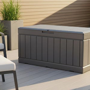 May include: A large, rectangular outdoor storage chest in shades of gray and brown. The chest has a slatted lid and vertical paneling on the sides. It is placed on a wooden deck, with a chair and greenery in the background.