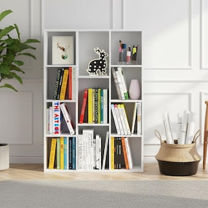 May include: A white bookshelf filled with books of various colors and sizes, decorative items, and office supplies. A large potted plant and a woven basket with rolled papers are also present. The room has a neutral color palette.