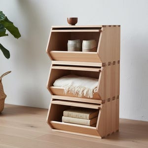 May include: A three-tiered wooden storage unit with a natural finish. The top shelf holds a small wooden bowl and two ceramic containers. The middle shelf contains a folded white blanket. The bottom shelf holds two books.
