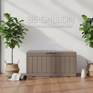 May include: A large, taupe-colored storage chest with a hinged lid, providing 85 gallons of space. The text on the wall reads "85 GALLON" and "Provide enough storage space for tools". Two potted plants flank the chest, with a rolled yoga mat in front.