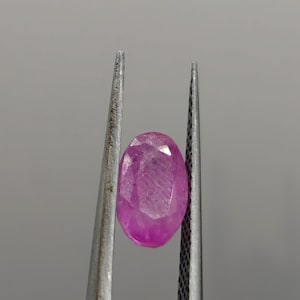 May include: A faceted, oval-shaped, pink gemstone is held by a pair of silver tweezers against a gray background. The gemstone has a polished surface and a vibrant, saturated color.