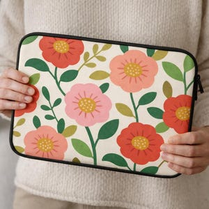 May include: A floral laptop sleeve featuring a cream background with a pattern of red, pink, and yellow flowers, green leaves, and black trim. The sleeve is held by a person.