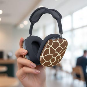 Puede incluir: Auriculares circumaurales de color gris oscuro con estampado de jirafa en los auriculares. Los auriculares tienen una diadema y almohadillas acolchadas. El diseño presenta un patrón de jirafa marrón y beige.